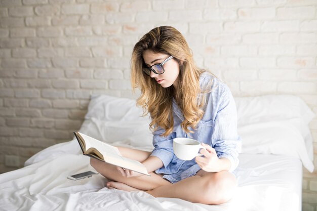 Portrait of a gorgeous young nerdy girl with glasses reading a book and drinking coffee in bed