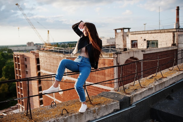 Free photo portrait of a gorgeous young woman in black leather jacket jeans and sneakers sitting on handrails on the roof with picturesque view of a park