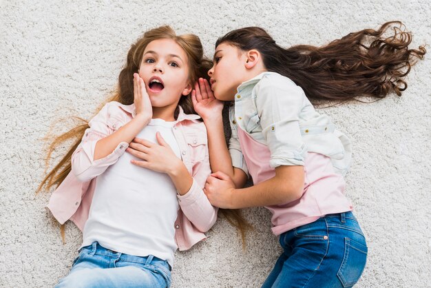 Portrait of a gossip girl telling a secret in the ear to her friend lying on carpet