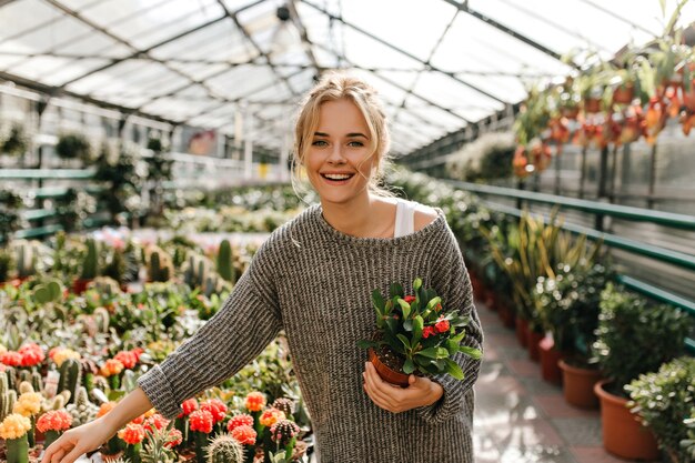 Portrait of green-eyed young woman holding plant. Woman in gray sweater walks in greenhouse.