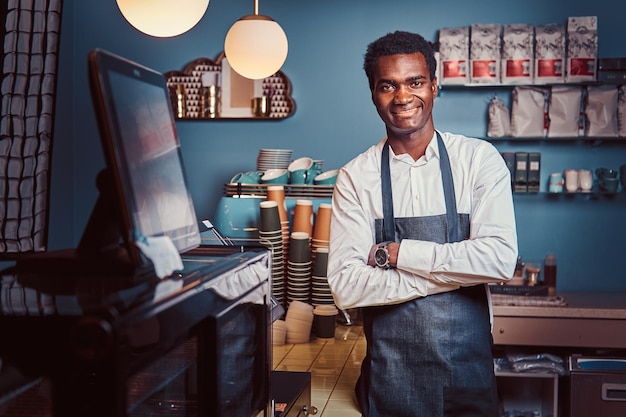 Portrait of a handsome African barista standing with crossed arms at the counter of a trendy coffee shop.