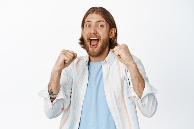 Portrait of handsome blond man cheering, scream yes, winning prize, making money on bet, rooting for sports team and shouting chanting, standing against white background.