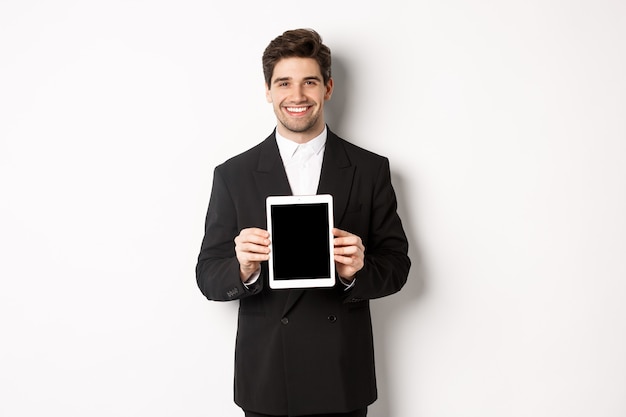 Portrait of handsome businessman in trendy suit, showing digital tablet screen and smiling, standing against white background