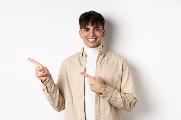 Portrait of handsome caucasian young man in glasses showing logo, smiling and pointing fingers left, inviting to check out promo deal, white background.