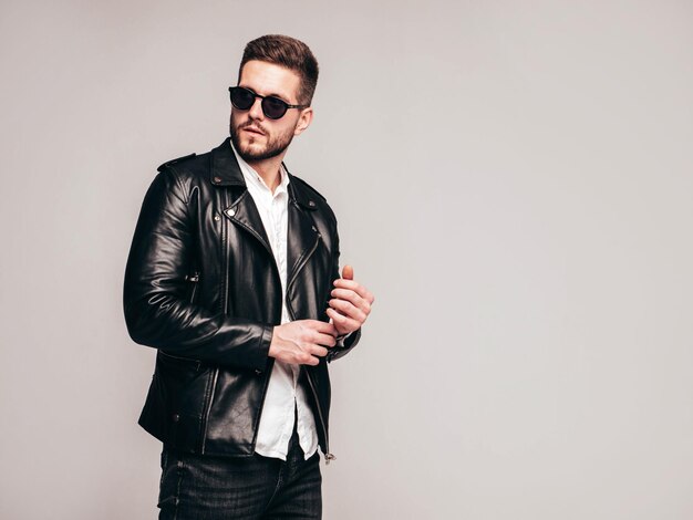 Portrait of handsome confident model Sexy stylish man dressed in biker leather jacket and black jeans Fashion hipster male isolated on grey background in studio in sunglasses Isolated