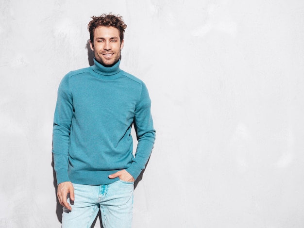 Free photo portrait of handsome confident model sexy stylish man dressed in blue sweater and jeans fashion hipster male with curly hairstyle posing near grey wall in studio isolated