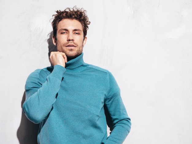 Free photo portrait of handsome confident model sexy stylish man dressed in blue sweater and jeans fashion hipster male with curly hairstyle posing near grey wall in studio isolated