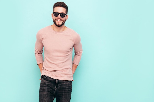 Portrait of handsome confident model Sexy stylish man dressed in pink sweater and jeans Fashion hipster male posing near blue wall in studio In sunglasses Isolated