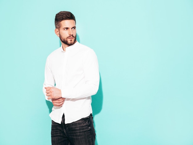 Free photo portrait of handsome confident model sexy stylish man dressed in white shirt and jeans fashion hipster male posing near blue wall in studio isolated