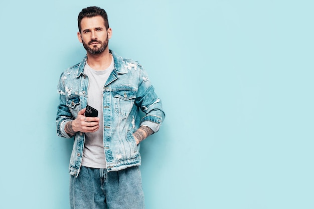 Portrait of handsome confident stylish hipster lambersexual model Sexy man dressed in jeans jacket Fashion male isolated on blue wall in studio Holding smartphone