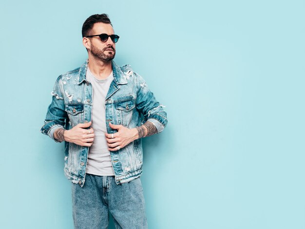 Portrait of handsome confident stylish hipster lambersexual model Sexy man dressed in jeans jacket Fashion male isolated on blue wall in studio In sunglasses