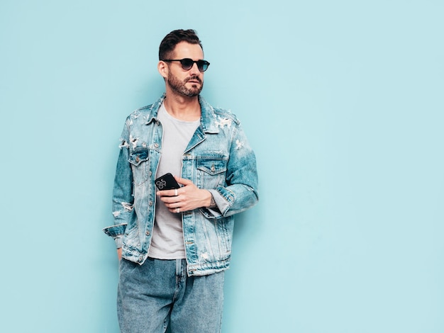 Portrait of handsome confident stylish hipster lambersexual model Sexy man dressed in jeans jacket Fashion male isolated on blue wall in studio In sunglasses