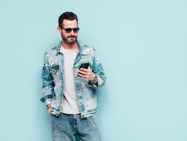 Portrait of handsome confident stylish hipster lambersexual model Sexy man dressed in jeans jacket Fashion male isolated on blue wall in studio In sunglasses