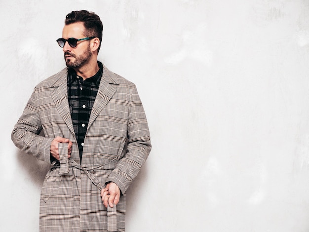 Portrait of handsome confident stylish hipster lambersexual model Sexy man dressed in jeans and overcoat Fashion male posing near grey wall in studio