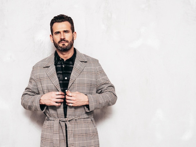 Portrait of handsome confident stylish hipster lambersexual model Sexy man dressed in jeans and overcoat Fashion male posing near grey wall in studio