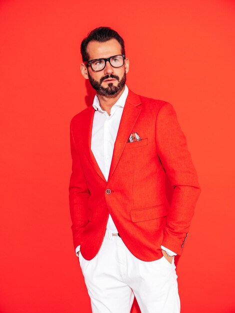 Portrait of handsome confident stylish hipster lambersexual model Sexy modern man dressed in elegant suit Fashion male posing in studio near red wall in spectacles