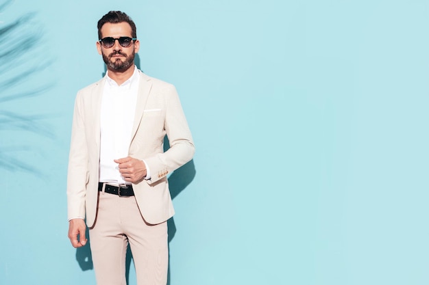 Portrait of handsome confident stylish hipster lambersexual model Sexy modern man dressed in white elegant suit Fashion male posing in studio near blue wall in sunglasses