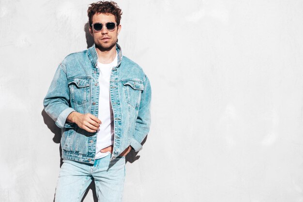 Portrait of handsome confident stylish hipster lambersexual model with curly hairstyle Sexy man dressed in jeans jacket Fashion male isolated on grey wall in studio In sunglasses