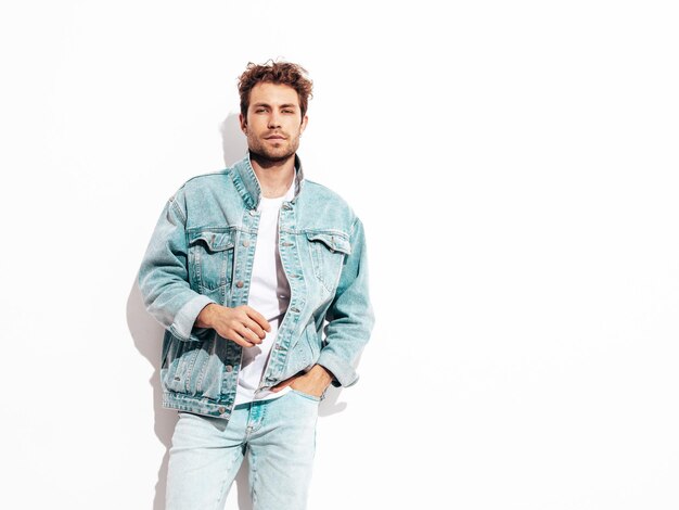 Portrait of handsome confident stylish hipster lambersexual model with curly hairstyle Sexy man dressed in jeans jacket Fashion male isolated on white wall in studio