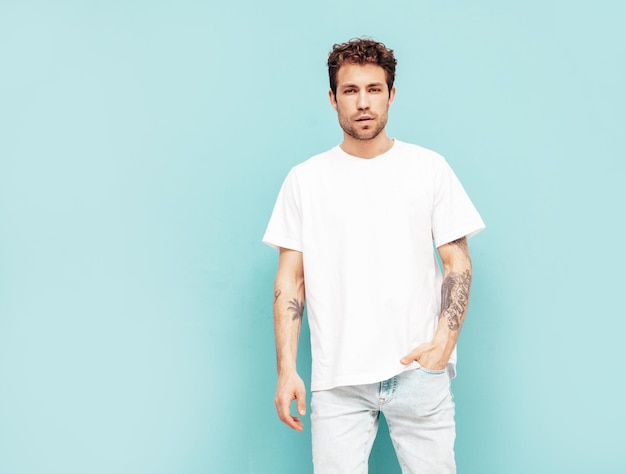 Free Photo portrait of handsome confident stylish hipster lambersexual model with curly hairstyle sexy man dressed in jeans and white tshirt fashion male isolated on blue wall in studio