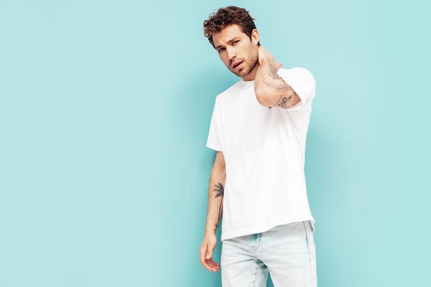 Free photo portrait of handsome confident stylish hipster lambersexual model with curly hairstyle sexy man dressed in jeans and white tshirt fashion male isolated on blue wall in studio