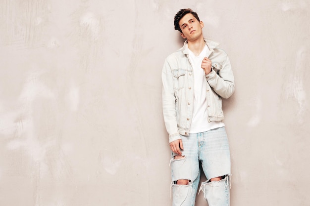 Portrait of handsome confident stylish hipster lambersexual modelMan dressed in jacket and jeans Fashion male posing in studio near grey wall