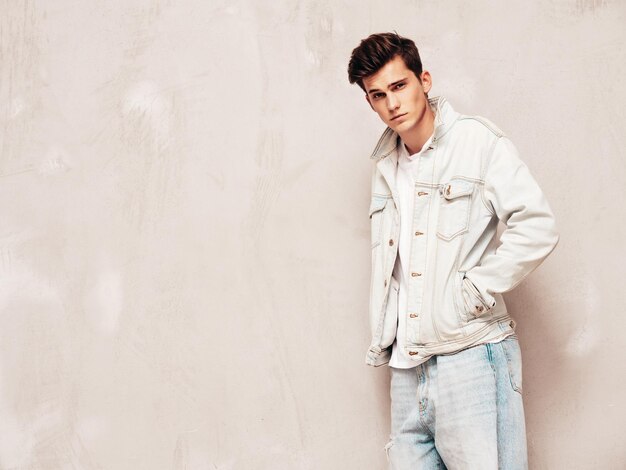 Portrait of handsome confident stylish hipster lambersexual modelMan dressed in jacket and jeans Fashion male posing in studio near grey wall