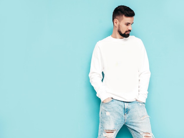 Free photo portrait of handsome confident stylish hipster lambersexual modelman dressed in white sweater and jeans fashion male isolated on blue wall in studio