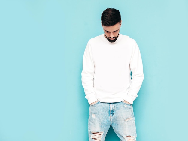 Portrait of handsome confident stylish hipster lambersexual modelMan dressed in white sweater and jeans Fashion male isolated on blue wall in studio