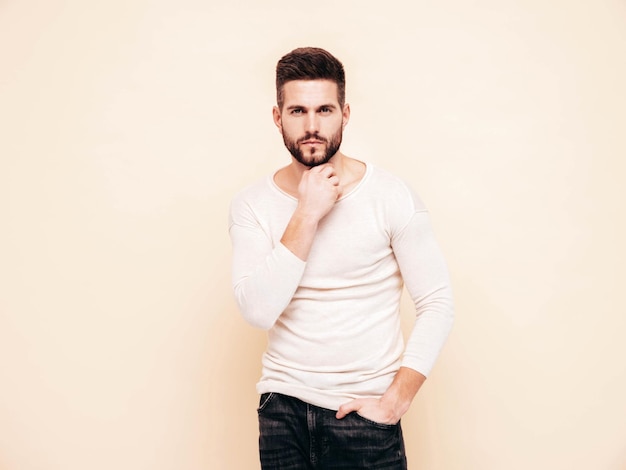 Portrait of handsome confident stylish hipster lambersexual modelMan dressed in white sweater and jeans Fashion male posing near wall in studio