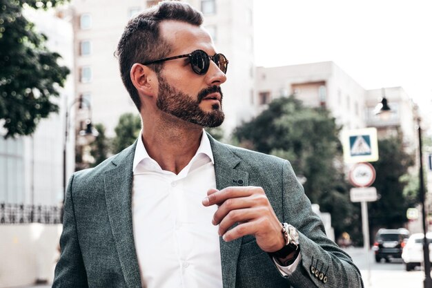 Portrait of handsome confident stylish hipster lambersexual modelModern man dressed in elegant suit Fashion male posing in the street background in Europe city at sunset In sunglasses