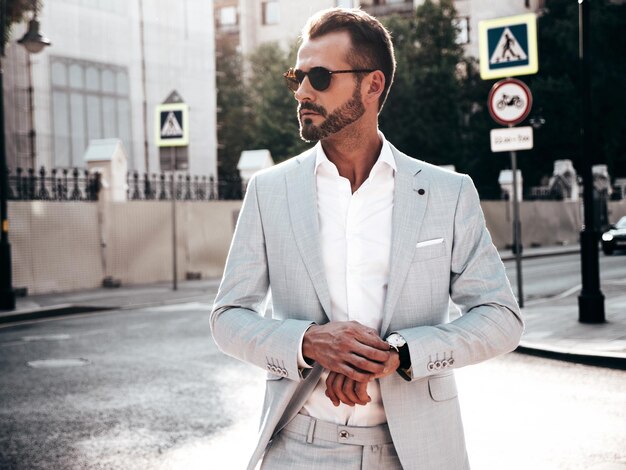 Portrait of handsome confident stylish hipster lambersexual modelModern man dressed in elegant white suit Fashion male posing in the street background in Europe city at sunset In sunglasses