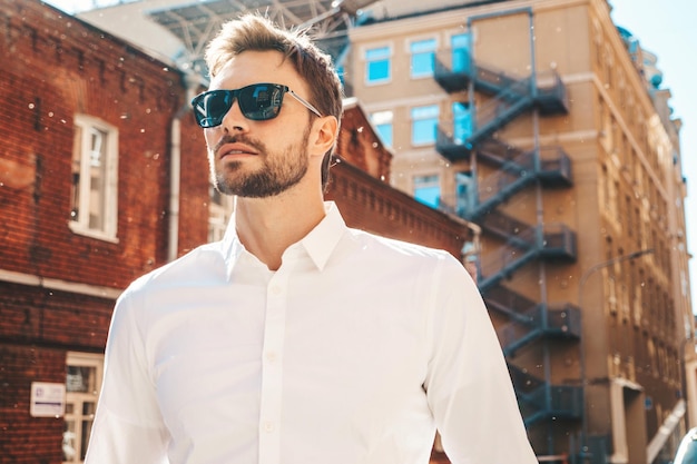 Free photo portrait of handsome confident stylish hipster lambersexual modelmodern man dressed in white shirt fashion male posing in the street background near skyscrapers in sunglasses outdoors at sunset