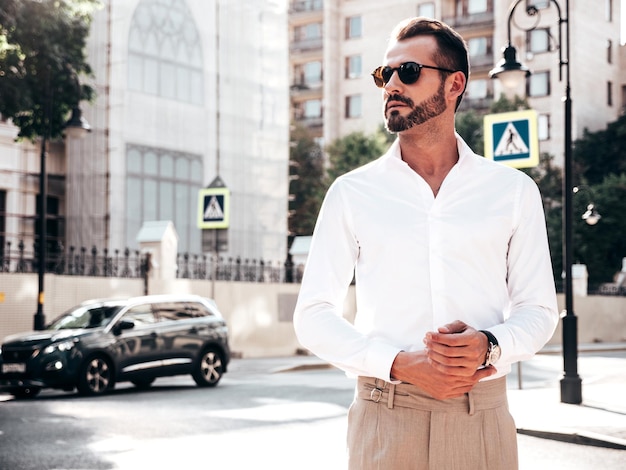 Portrait of handsome confident stylish hipster lambersexual modelModern man dressed in white shirt and trousers Fashion male posing in the street background in Europe city at sunset In sunglasses