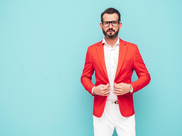 Portrait of handsome confident stylish hipster lambersexual modelSexy modern man dressed in elegant red suit Fashion male posing in studio near blue wall in spectacles