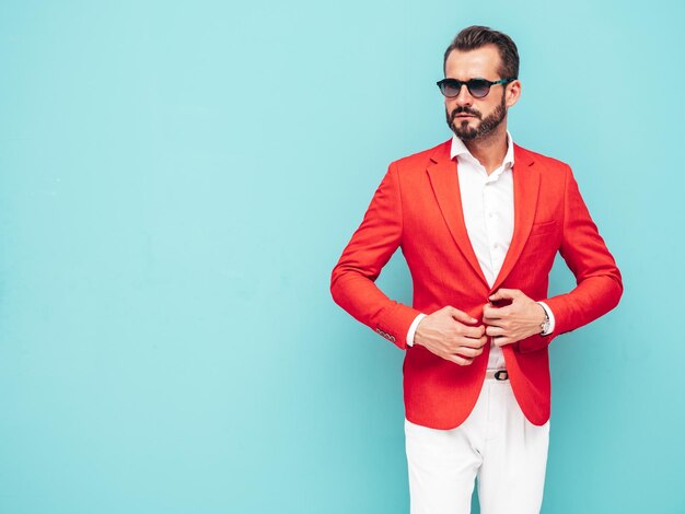 Portrait of handsome confident stylish hipster lambersexual modelSexy modern man dressed in elegant red suit Fashion male posing in studio near blue wall in sunglasses