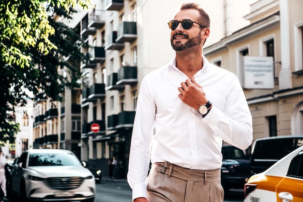 Free photo portrait of handsome confident stylish hipster lambersexual modelsexy modern man dressed in white shirt and trousers fashion male posing on street background in europe city at sunset in sunglasses