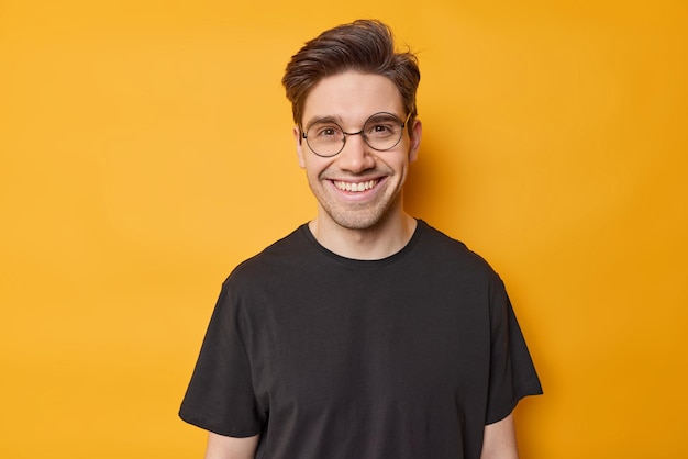 Free photo portrait of handsome dark haired man smiles toothily feels glad wears round spectacles and casual black t shirt expresses positive emotions isolated over yellow background happiness concept