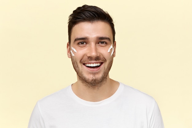 Portrait of handsome dark skinned unshaven male having energetic happy facial expression, looking at camera with broad radiant smile with stripes of white cream on cheeks