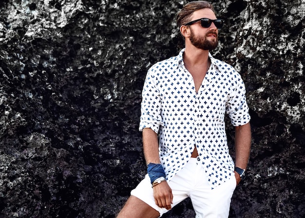 Portrait of handsome fashion man model wearing white clothes posing near rocks on the beach