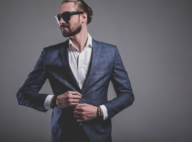 portrait of handsome fashion stylish hipster businessman model dressed in elegant blue suit in sunglasses posing on gray