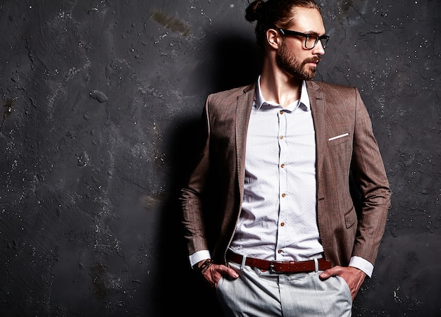 Free photo portrait of handsome fashion stylish hipster businessman model dressed in elegant brown suit in glasses near dark wall