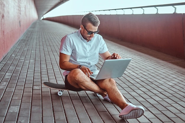 Free photo portrait of a handsome fashionable freelancer in sunglasses dressed in a white shirt and shorts working on a laptop while sitting on a skateboard under a bridge.