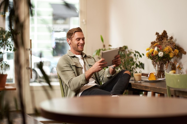 Free photo portrait of handsome happy guy young man sits in cafe watching videos on digital tablet wearing