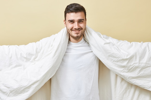 Portrait of handsome joyful young European huy with broad radiant smile holding large white blanket with his arms outstretched, going to sleep finally after hard working day