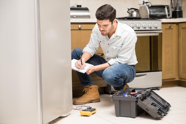 Free photo portrait of a handsome male technician doing a work report on a broken fridge in a home kitchen