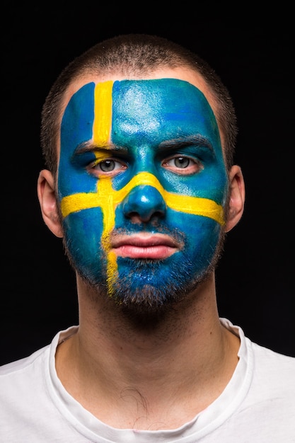 Free photo portrait of handsome man face supporter fan of sweden national team with painted flag face isolated on black background. fans emotions.