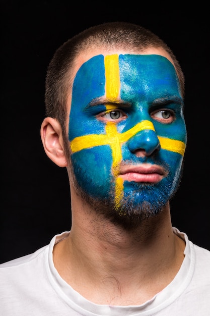 Free photo portrait of handsome man face supporter fan of sweden national team with painted flag face isolated on black background. fans emotions.