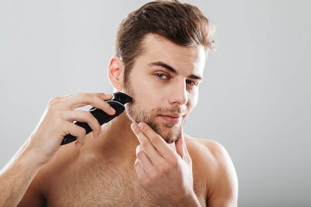 Portrait of handsome man shaving his beard with electric shaver in morning against grey wall