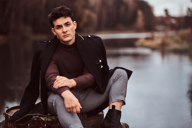 Portrait of a handsome man with a coat covering his shoulders looking at camera while sitting near a lake in the autumn forest.